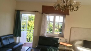 Een kamer met een zwarte leren bank, een groene bank, een kleine tafel en een bed. Er is een raam met gordijnen en een kroonluchter.