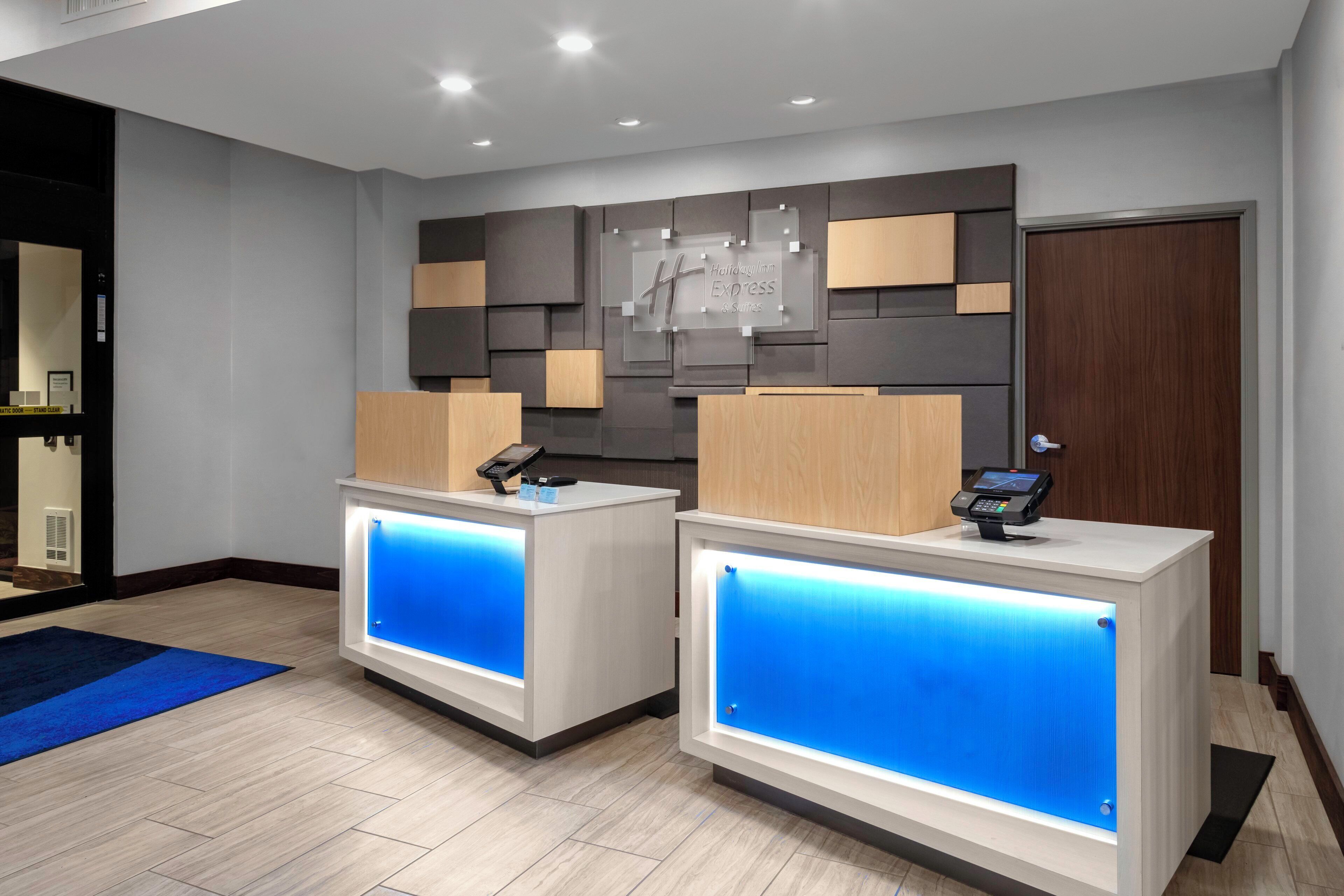 A hotel reception desk with a modern design, featuring two reception counters with illuminated blue panels, a digital display, and a wall-mounted sign above.