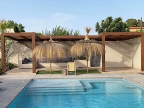 Une piscine agrémentée d’une pergola et d’un espace détente, entourée de verdure et surplombée par un ciel dégagé.