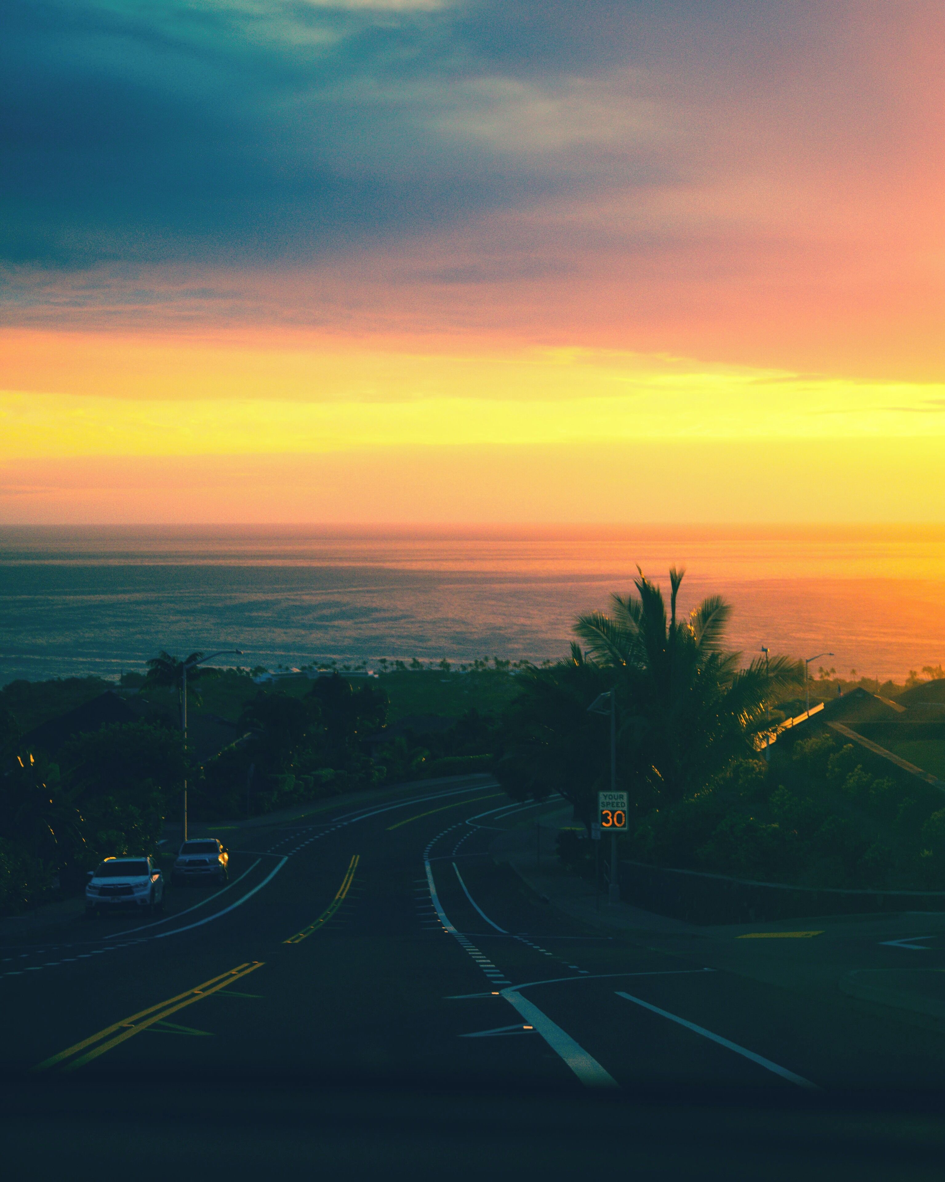 Una carretera que lleva hacia un atardecer, con palmeras y una señal de límite de velocidad.