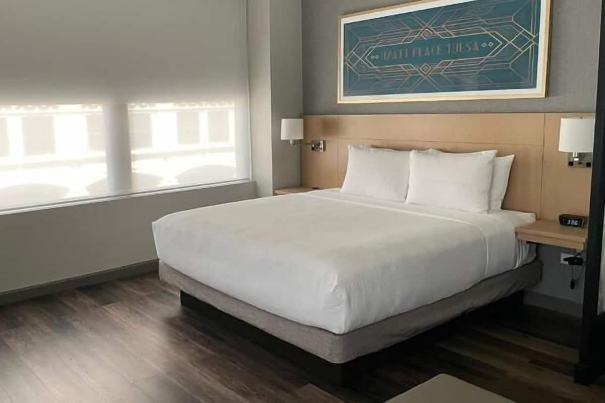 Ein Hotelzimmer mit einem großen Bett, einem Fenster mit Jalousien, einem Wandbild und einem Nachttisch mit Lampe.