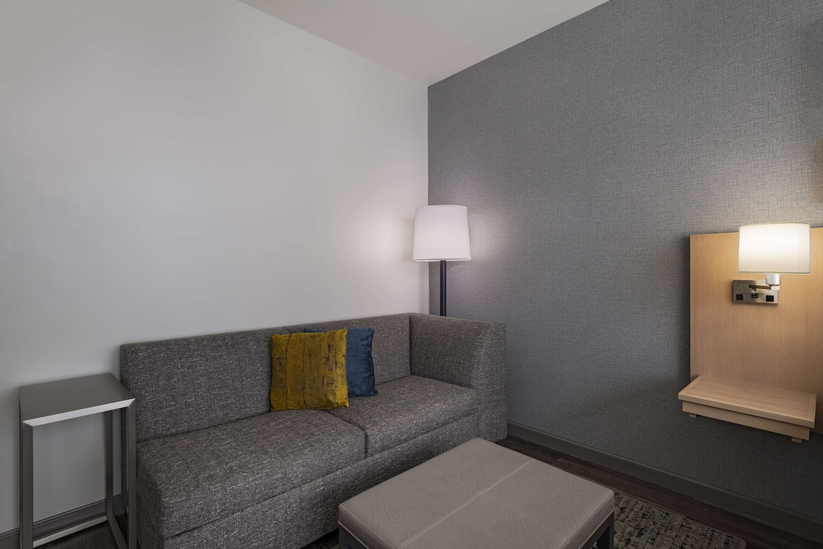 Ein modernes Hotelzimmer mit grauem Sofa, einem kleinen Beistelltisch, einer Lampe und einem Wandregal.