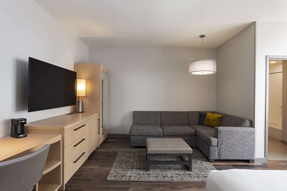 Ein modernes Hotelzimmer mit Flachbildfernseher, einem Holztisch, einem grauen Sofa, einem kleinen Hocker und einer Pendelleuchte.
