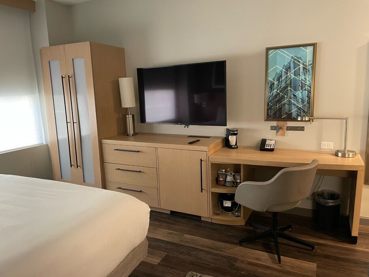 Quarto de hotel com cama, escrivaninha com cadeira, televisão, máquina de café e lixeira.