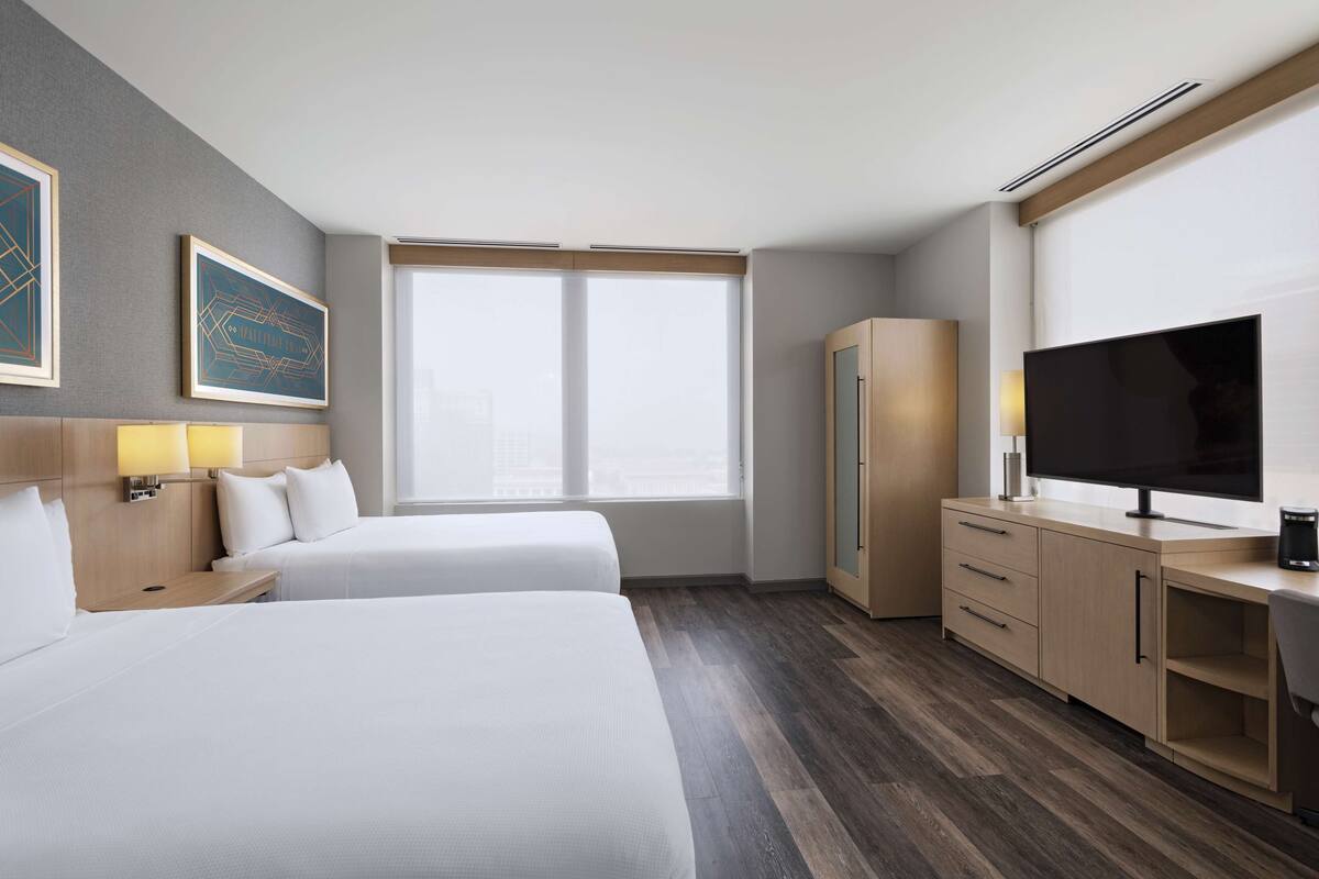 Ein Hotelzimmer mit zwei Betten, einem Schreibtisch mit Fernseher, einem Kleiderschrank und einem großen Fenster.