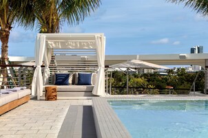 Zona de piscina con un toldo blanco, un área de descanso acolchada con cojines azules y una palmera.