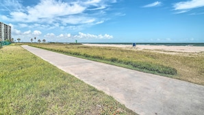Ein Betonweg führt zu einem Strand mit blauem Himmel und Palmen in der Ferne.