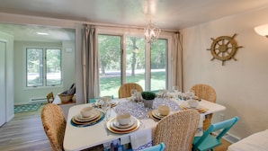 A dining area with a round table set for six, surrounded by wicker chairs, a chandelier, and a ship's wheel decoration on the wall. - Updated Home w/ Deck: 2 Mi to Sister Bay Beach! (Sister Bay)