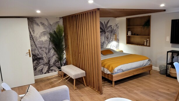 A bedroom with a bed, a wooden partition, a bench, a shelf with bottles, and a wall mural.