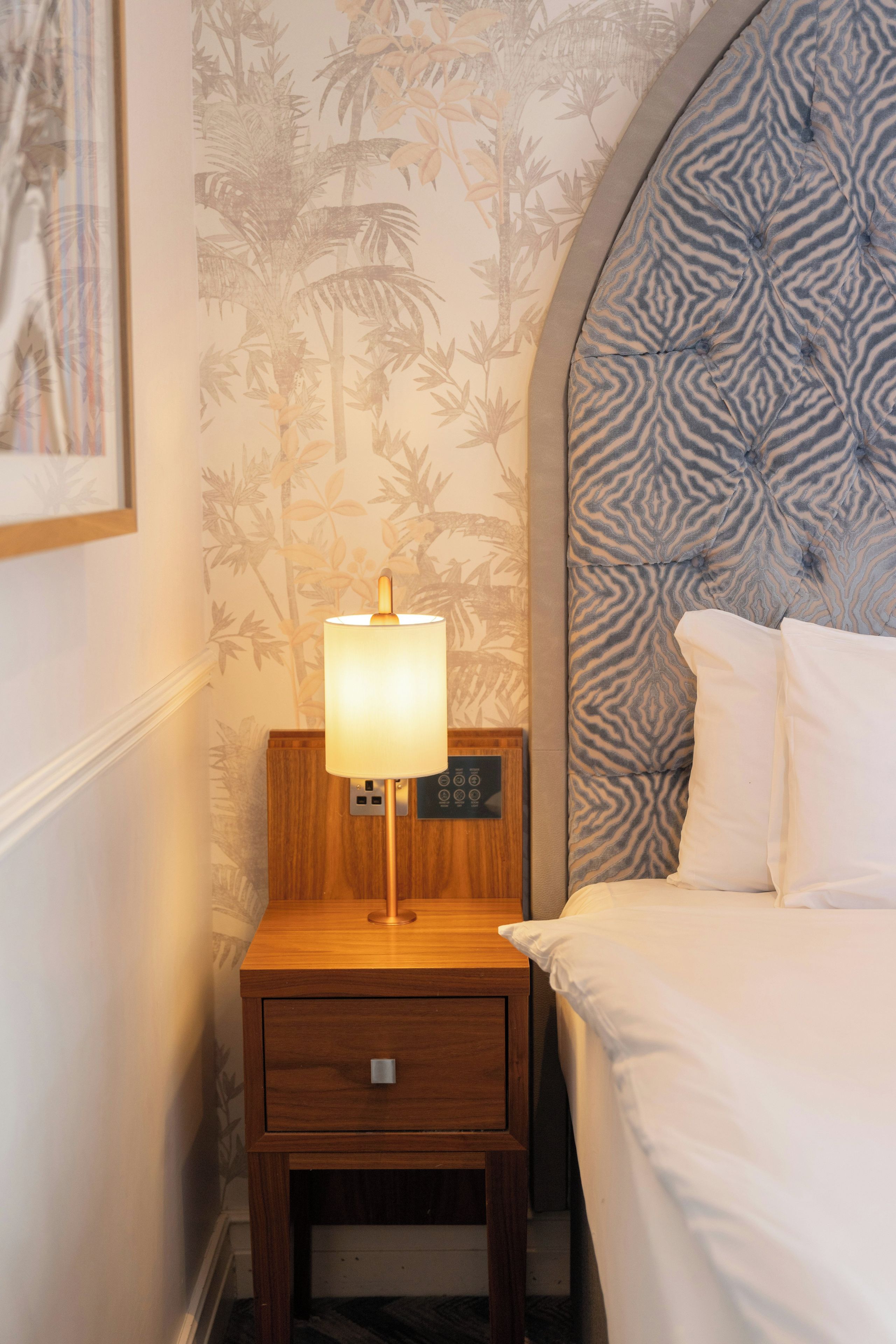 A hotel room with a bed, a nightstand with a lamp, and a wall with a tropical wallpaper pattern.