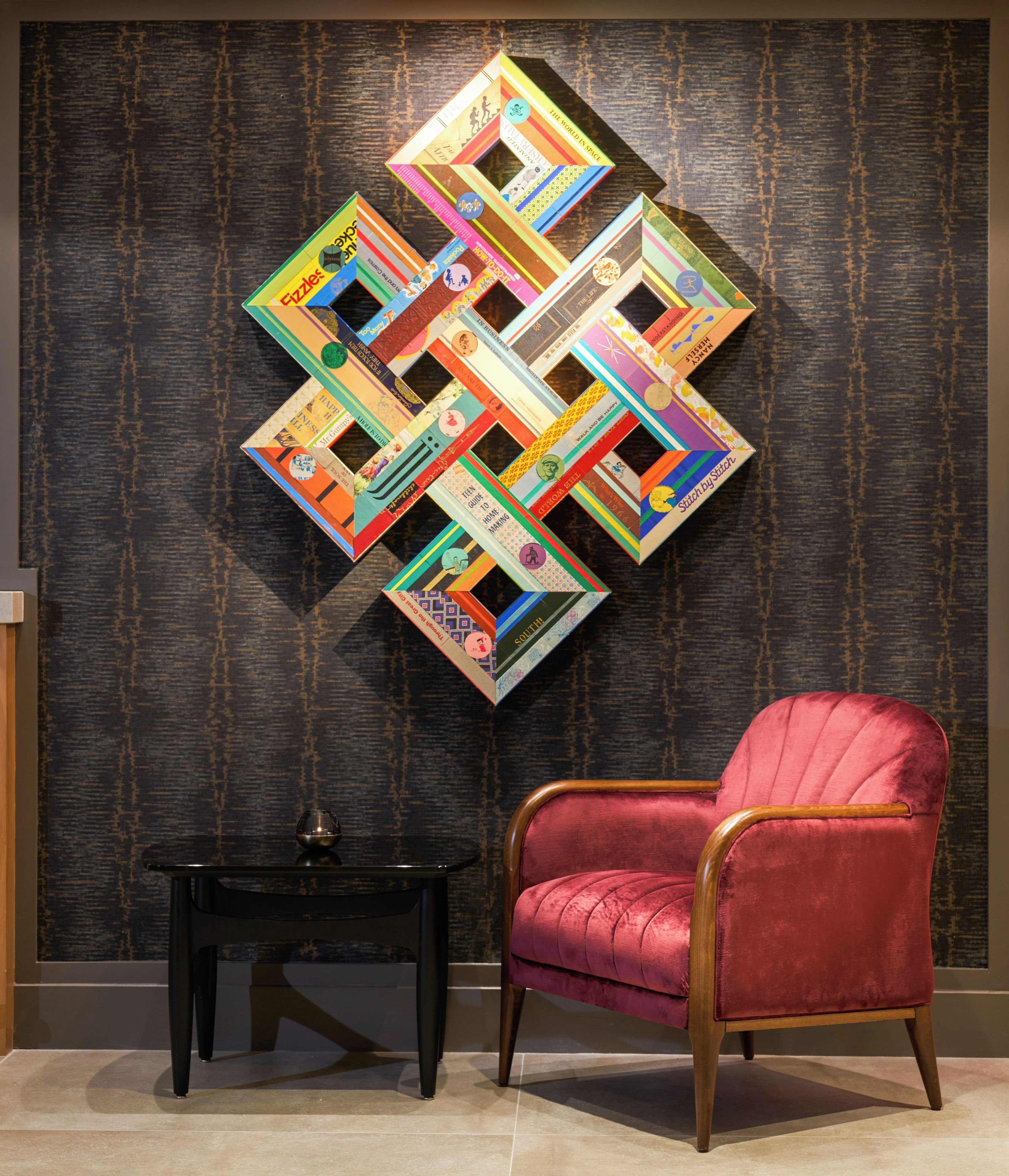 A red velvet armchair and a black side table in a room with a textured wall and a colorful geometric wall art piece.