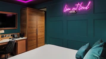 Quarto de hotel com parede de destaque verde-água, um letreiro de neon com a inscrição 'Love on the Road', um guarda-roupa de madeira, uma escrivaninha com um computador e uma cama com travesseiros verde-água.