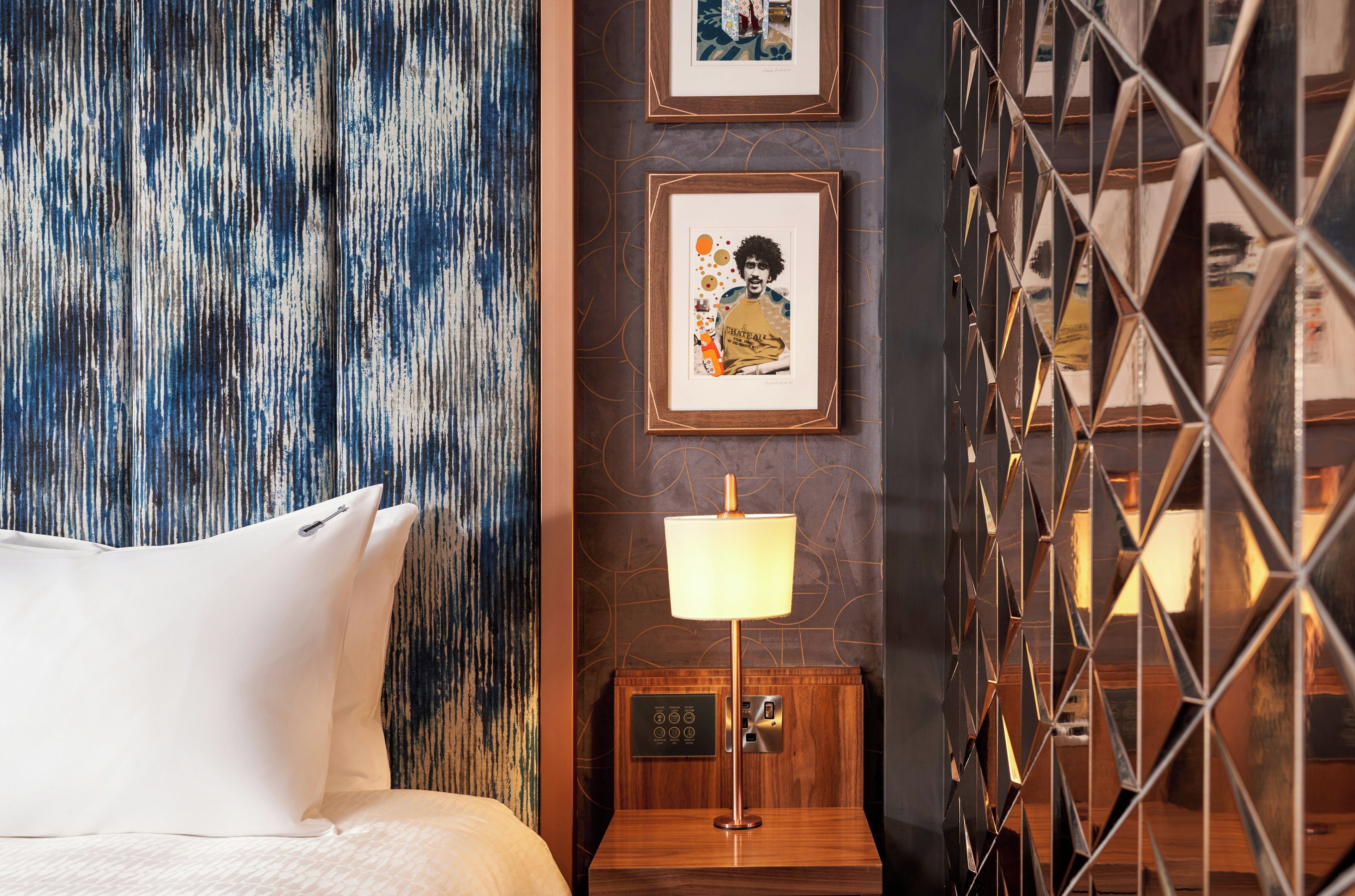 Una habitación de hotel con una cama, una pared con un diseño abstracto azul, un cuadro enmarcado, una lámpara de pie y un separador geométrico de metal.