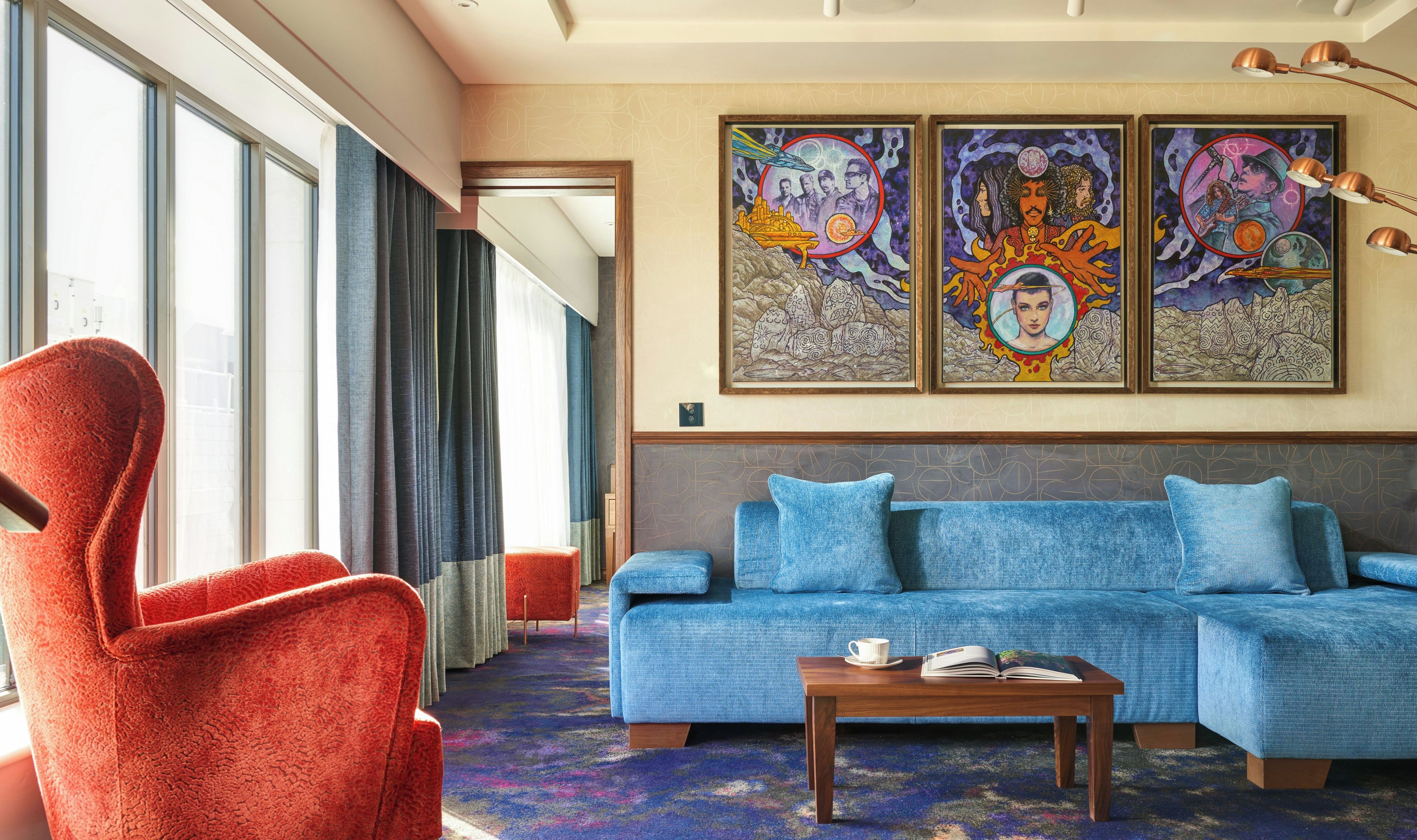A modern living room with a large window, a red armchair, a blue sofa, and two framed artworks on the wall.