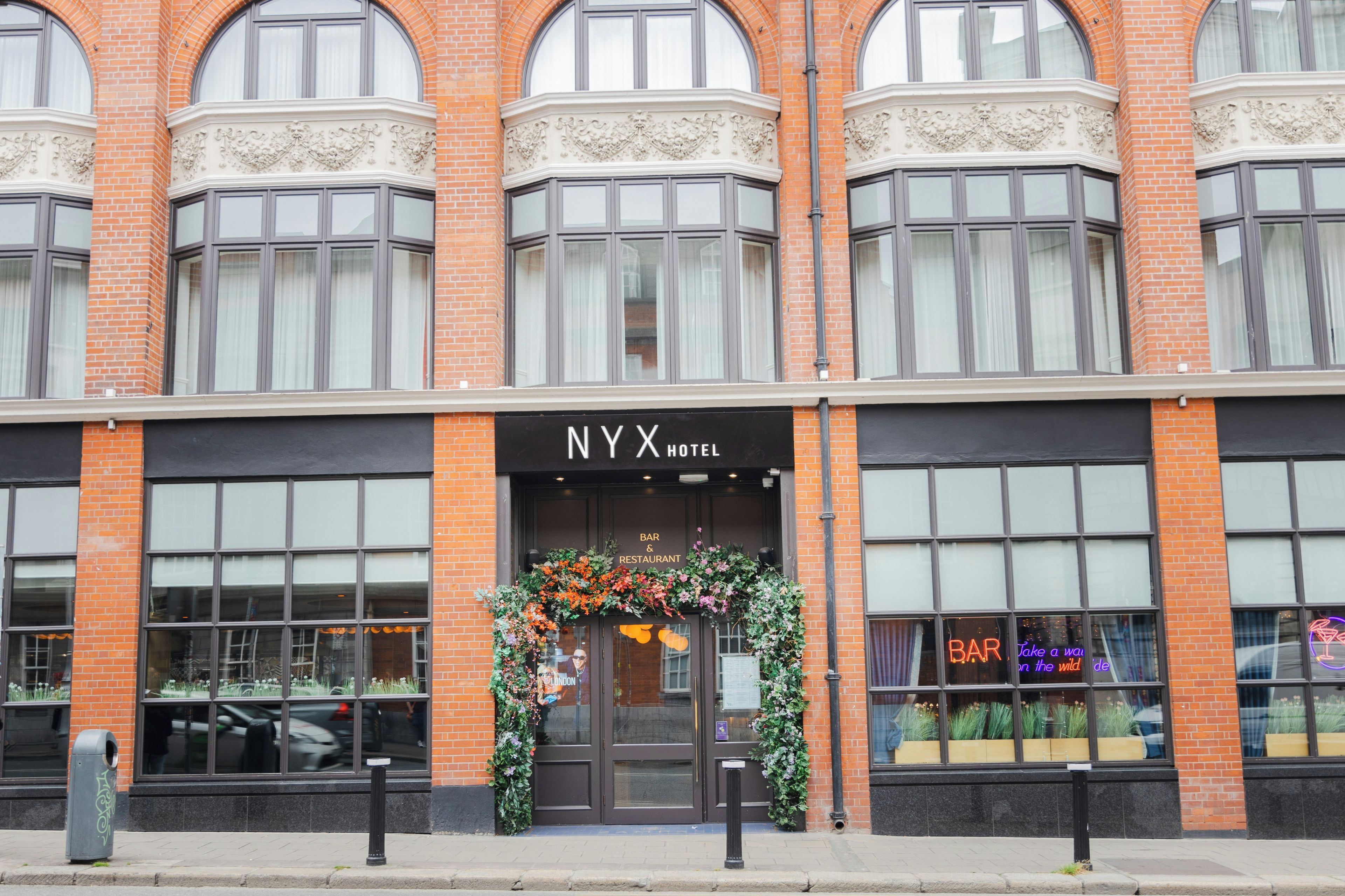 A hotel entrance with a black awning, a sign reading 'NYX HOTEL', and a floral decoration above the door.