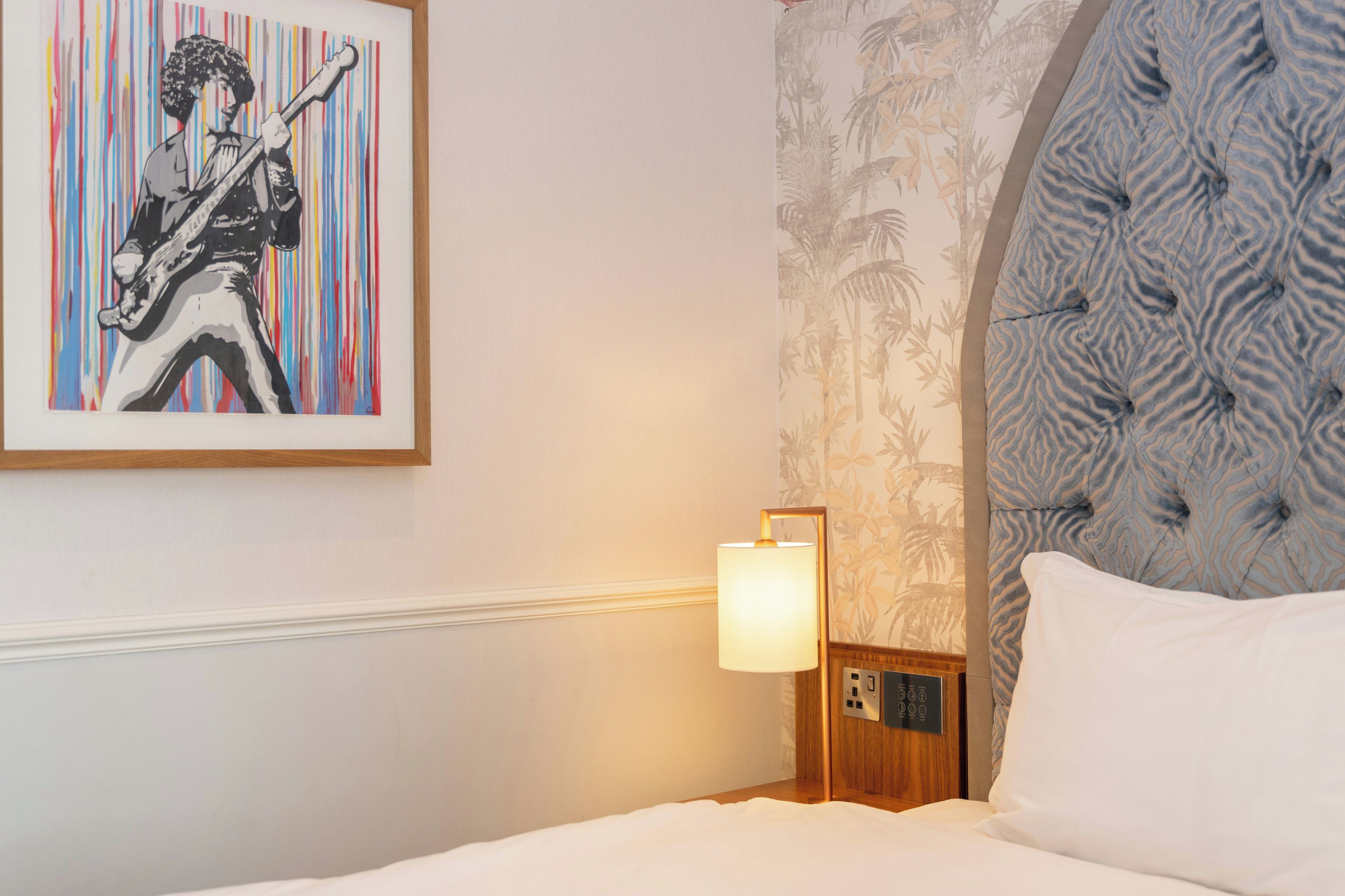 A hotel room with a bed, a nightstand with a lamp, a wall with a framed artwork, and a patterned headboard.