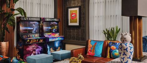 Un vestíbulo de hotel con juegos de arcade, un sofá con cojines de colores, un jarrón decorativo y una planta grande.