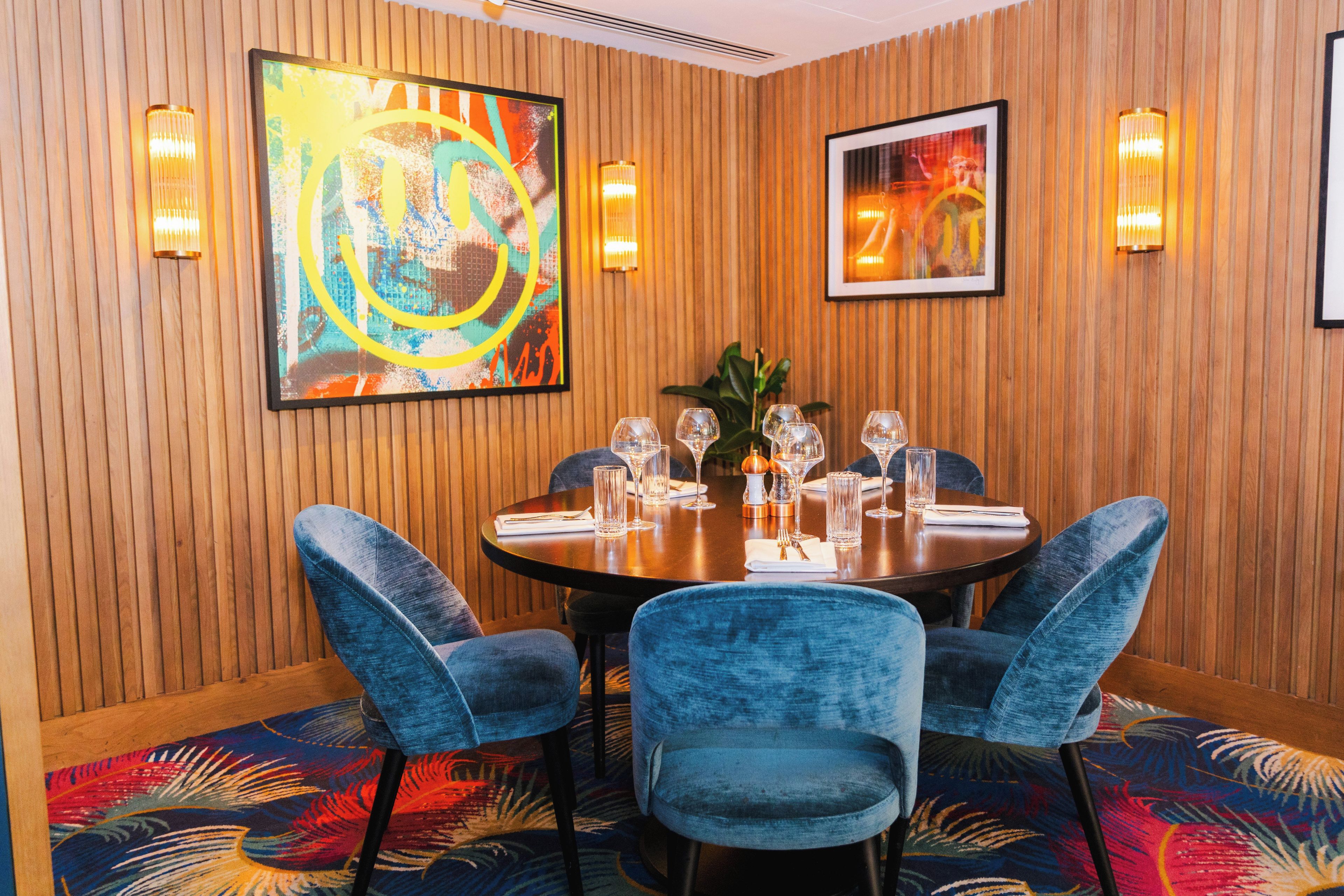 A dining area with a round table, four blue upholstered chairs, and two framed abstract paintings on the wall.