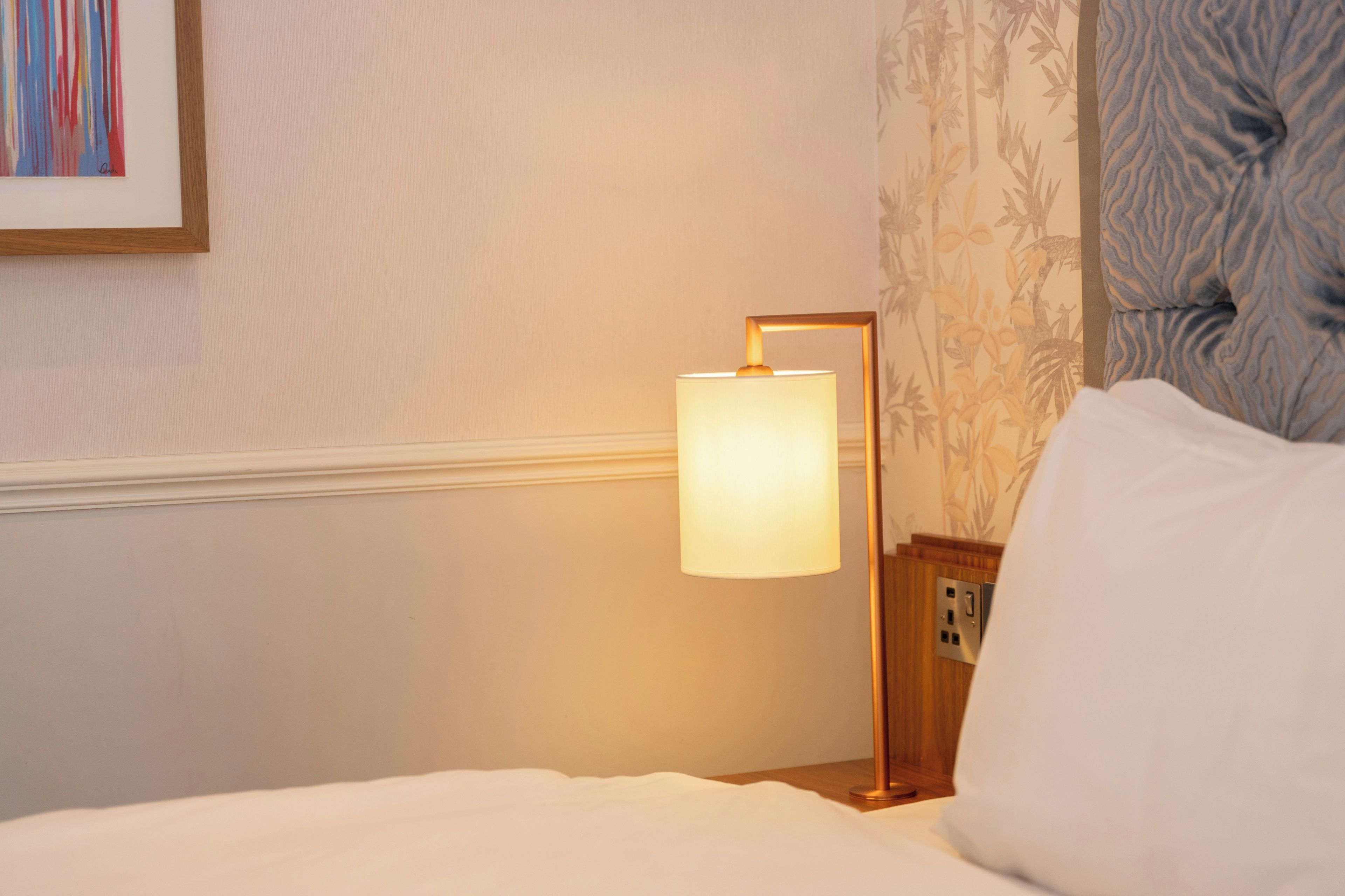 A hotel room with a bed, a bedside table with a lamp, and a wall with a framed picture.