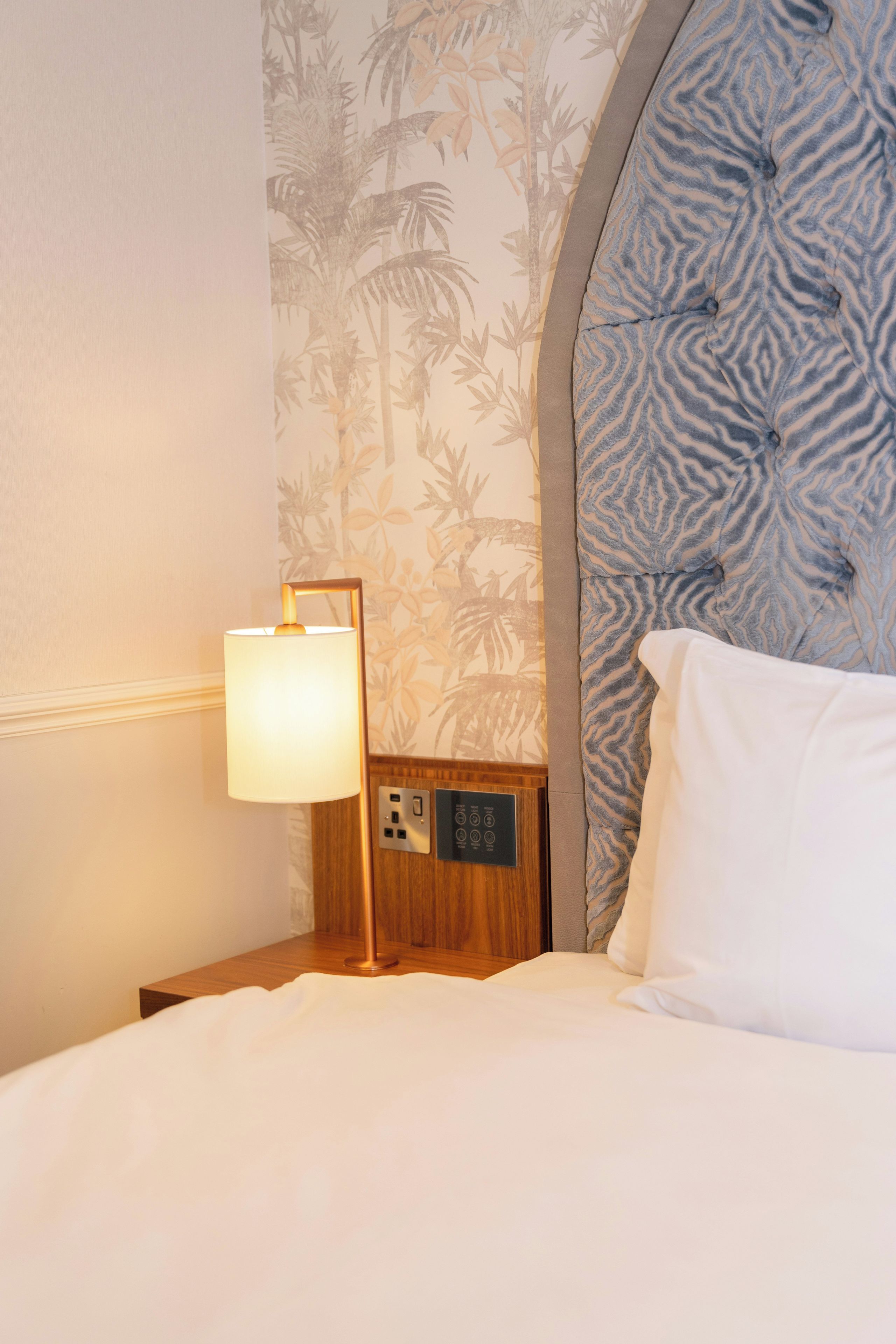 A hotel room with a bed, a nightstand, a lamp, and a wall with a tropical wallpaper pattern.