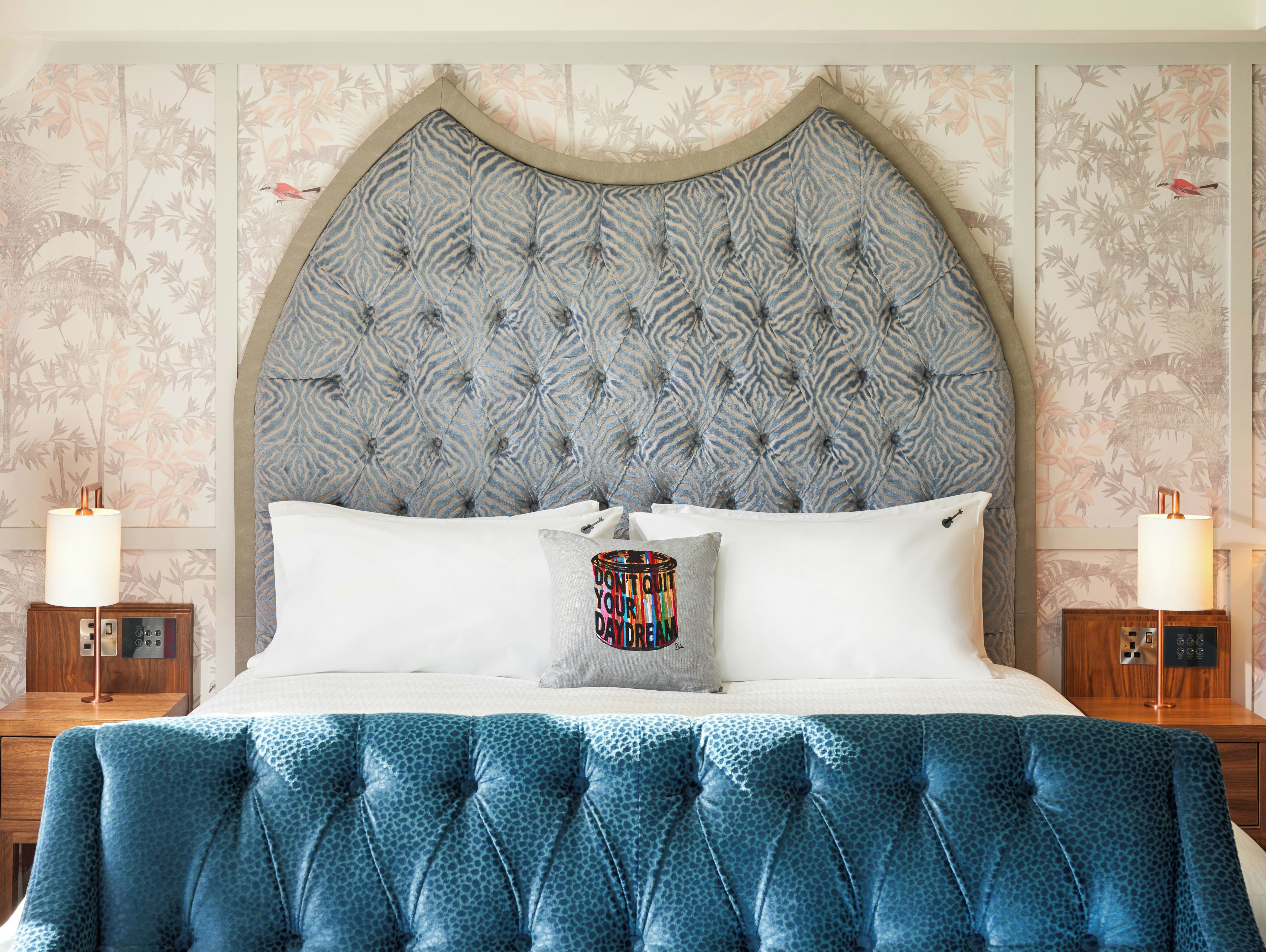 A bedroom with a tufted headboard, a bed with white linens, a blue patterned bedspread, and two bedside tables with lamps.