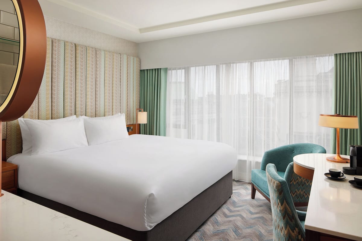 A hotel room with a large bed, a desk with a lamp, a chair, and a view of the city through sheer curtains.