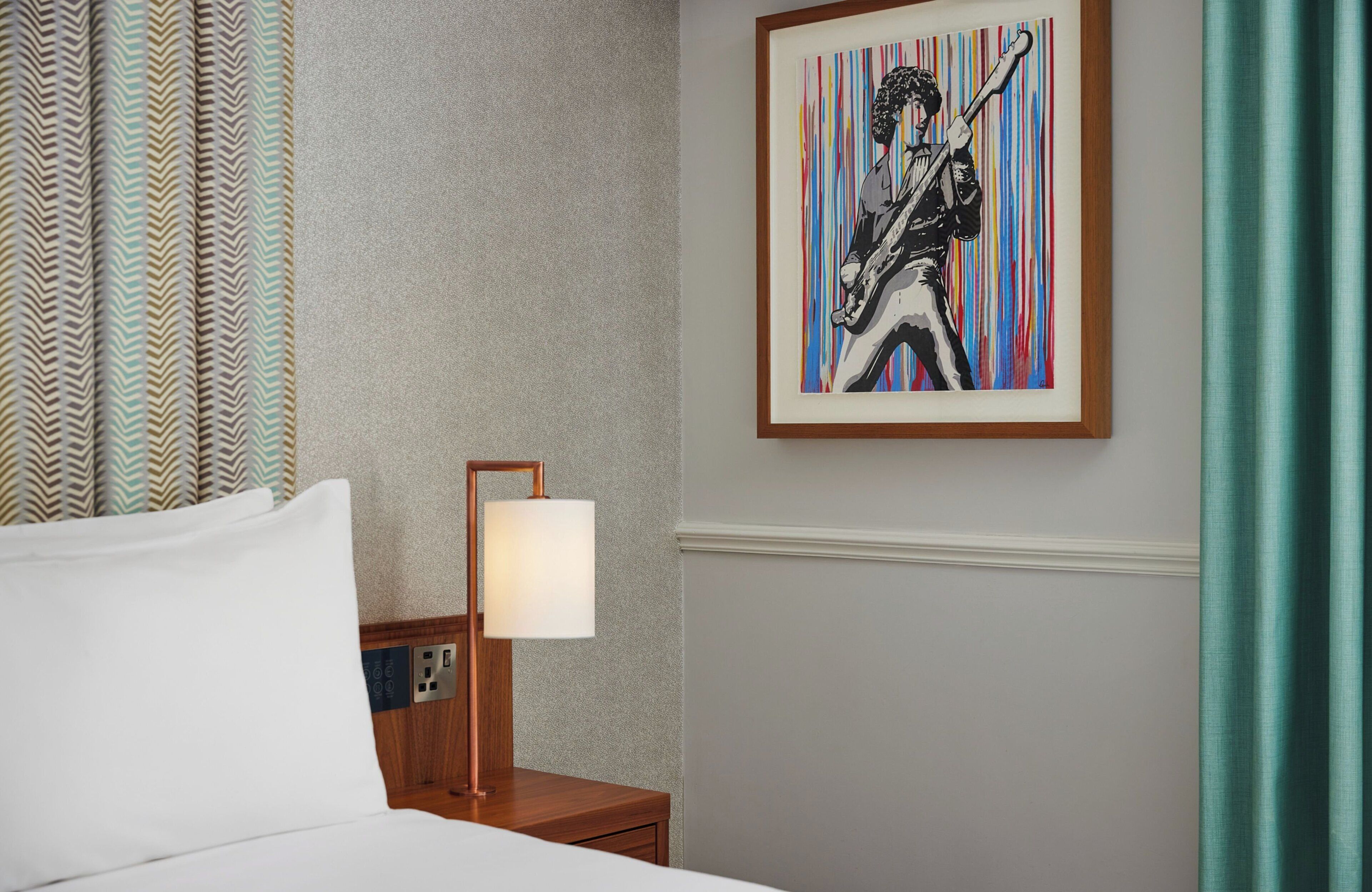 Habitación de hotel con cama, mesita de noche con lámpara, una obra de arte enmarcada en la pared y cortinas estampadas.