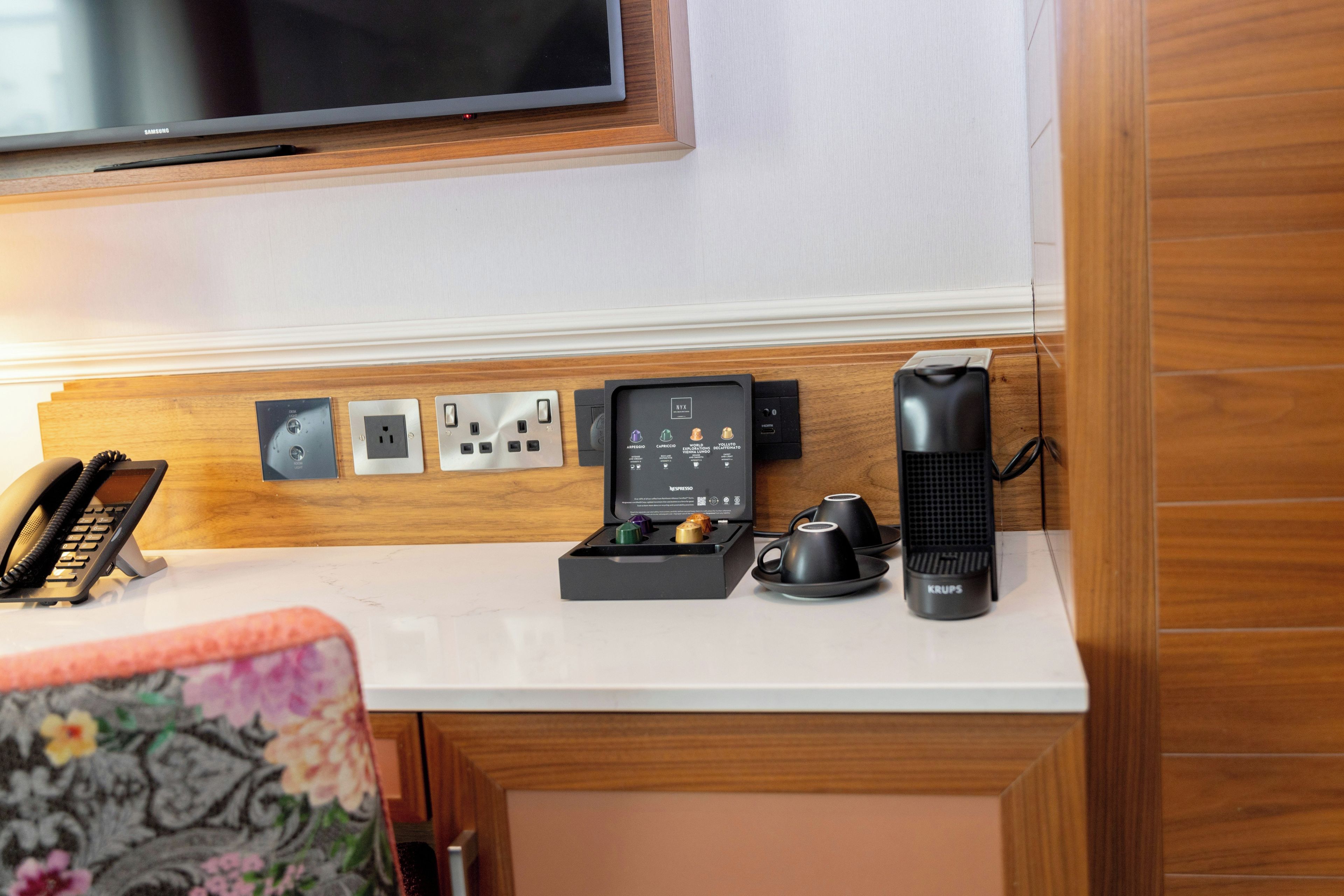 Una kitchenette con un televisor Samsung montado sobre una encimera blanca, un teléfono, una cafetera y una máquina de café espresso Krups.