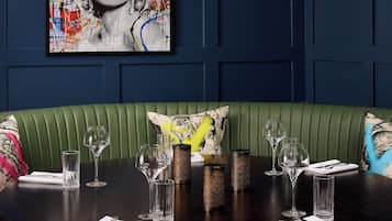 Un comedor con una mesa redonda dispuesta para cuatro personas, asientos tipo banca tapizados en verde y un cuadro abstracto grande en la pared.