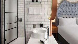 Un baño moderno con cabina de ducha de vidrio, un lavabo blanco y un espejo redondo, y una cama con cabecera capitonada.