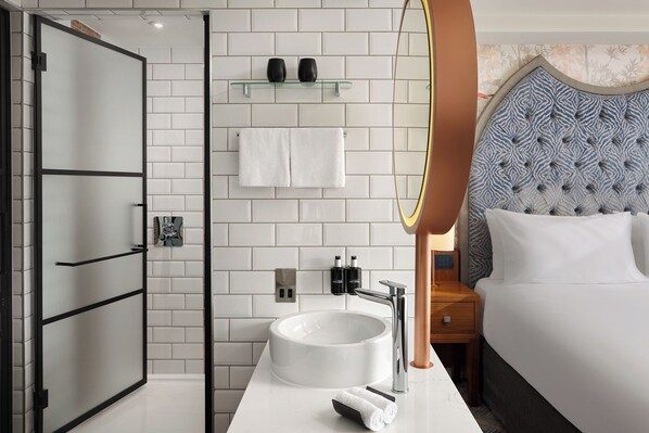 Un baño moderno con cabina de ducha de vidrio, un lavabo blanco y un espejo redondo, y una cama con cabecera capitonada.