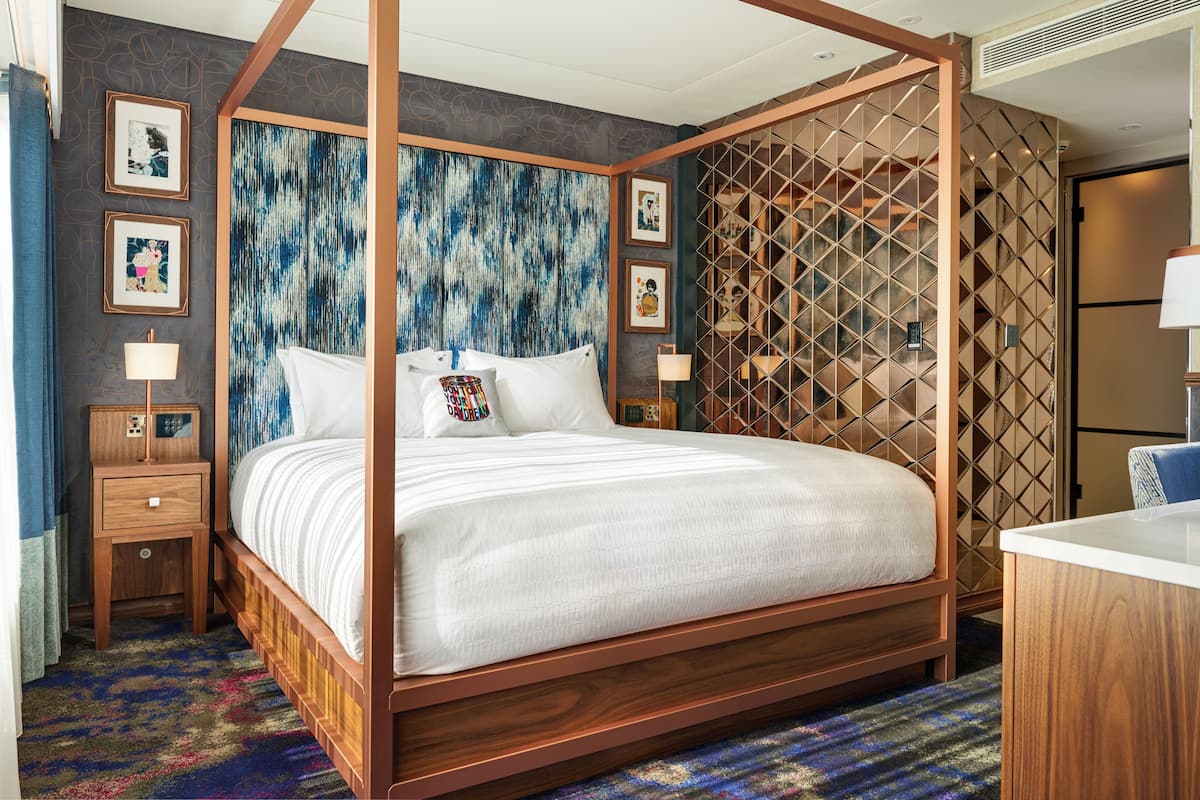 A four-poster bed with a wooden frame, a patterned wall behind it, and a small bedside table with a lamp.