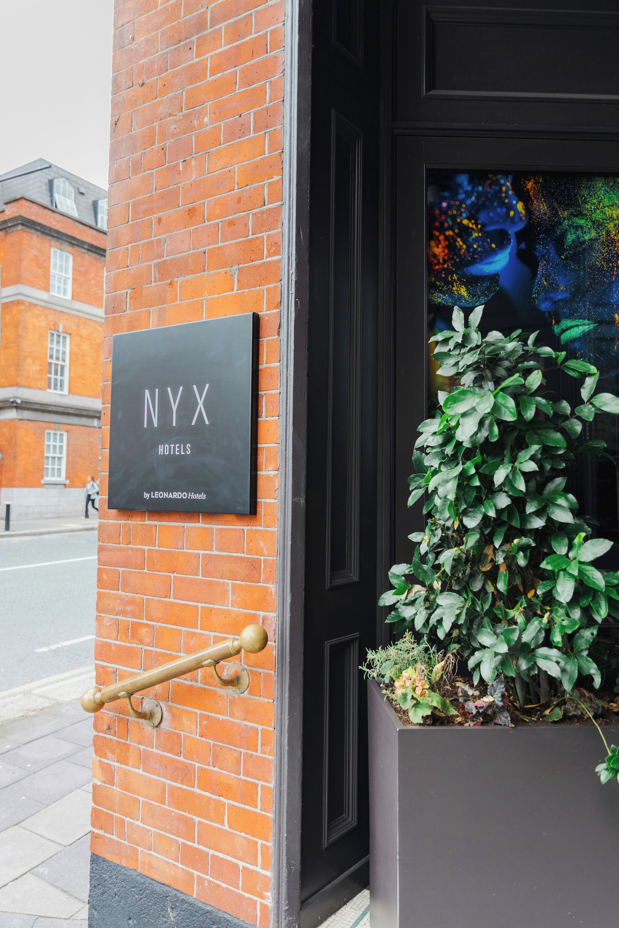 A NYX Hotels sign on a brick wall, a black door, and a potted plant with green foliage.