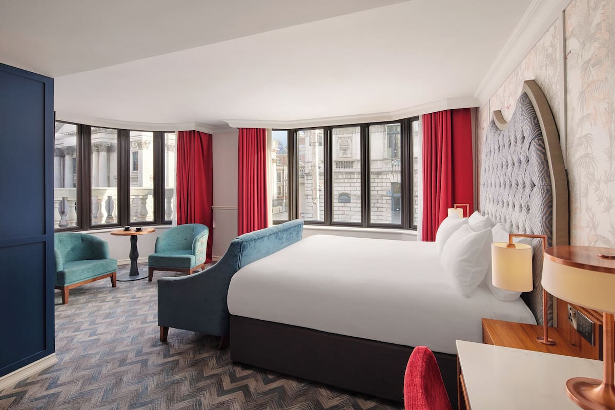 A hotel room with a large bed, two armchairs, a small table, and a view of the city through large windows.