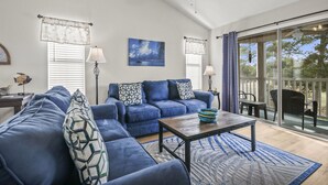A living room with a blue sofa set, a coffee table, and a view of the outdoors through large glass doors. - Pawleys Island Condo with Golf Views  Minutes to beach Pool/Hot Tub Dog-friendly (Pawleys Island)