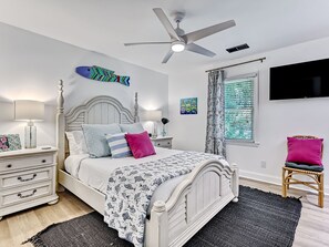 A bedroom with a white bed, a ceiling fan, a TV mounted on the wall, and a window with curtains. - Heated Pool Access | 5*Clean | Flex Cancelation (Tybee Island)