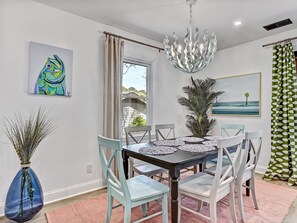 A dining area with a table, chairs, a chandelier, artwork, and a view through the window. - Heated Pool Access | 5*Clean | Flex Cancelation (Tybee Island)