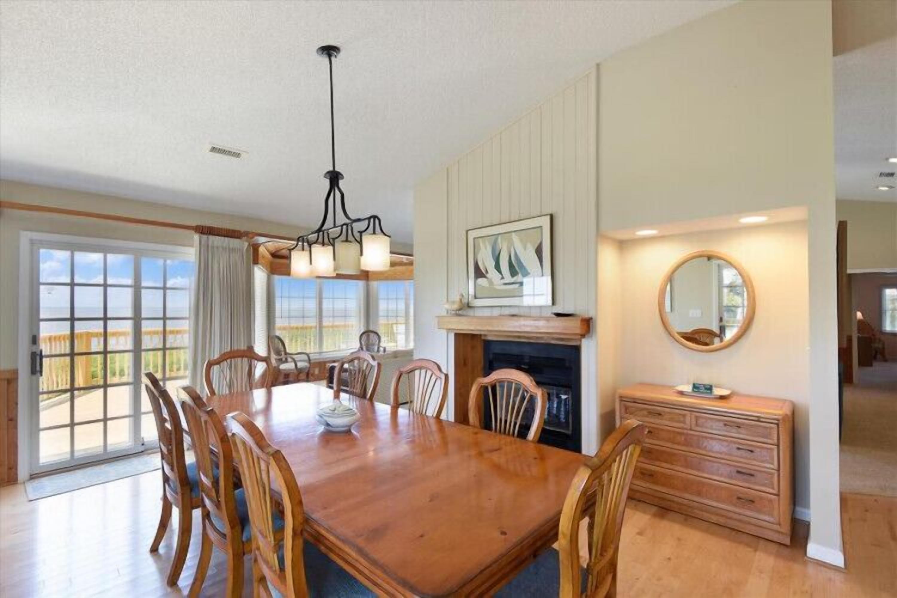 A dining room with a wooden table, chairs, a chandelier, a fireplace, and a large window with a view.