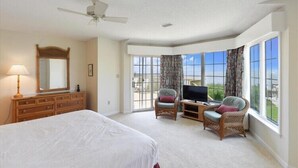 A hotel room with a large bed, a television, two wicker chairs, a dresser, and a balcony with curtains. - The Tayer-Soundfront at Ships Watch in Duck (Duck)