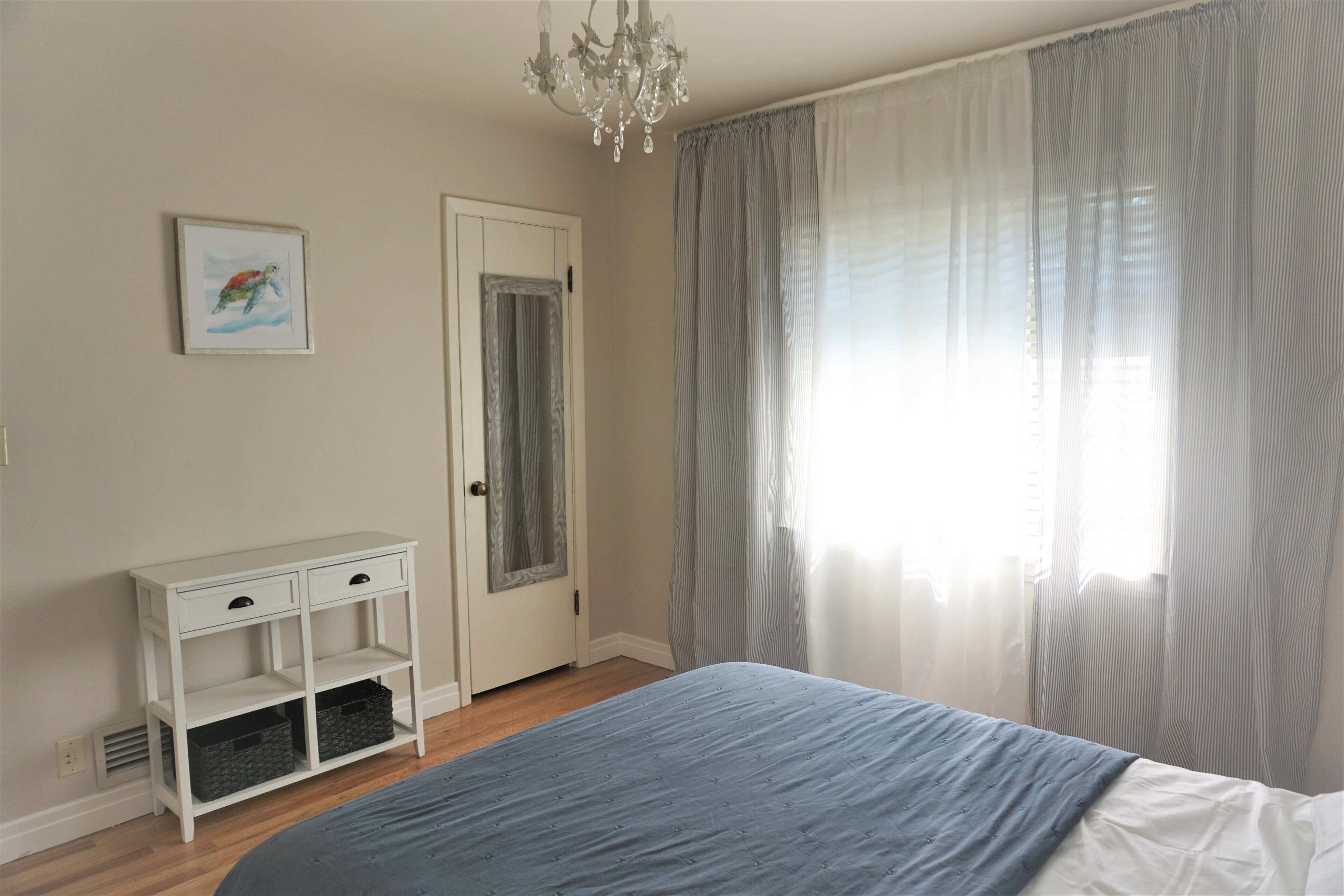Un dormitorio con una cama, una mesita de noche con cajones, un estante pequeño, una puerta y una ventana con cortinas traslúcidas.