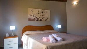 Un dormitorio con cabecera de madera, una cama con colcha color crema, dos lámparas de noche, una mesita de noche con lámpara, y un aplique de pared.