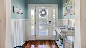 Um corredor com piso de madeira, uma porta branca adornada com uma guirlanda e uma pequena mesa com objetos decorativos.