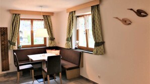 Una habitación pequeña y acogedora con un banco de madera, una mesa y dos sillas. Hay dos ventanas con cortinas, y un estante en la pared con adornos.