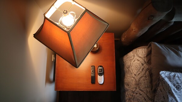 A wooden nightstand with a lamp, two remote controls, and a patterned pillow on a bed.
