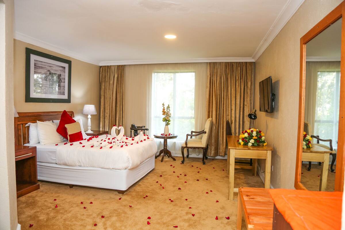 A hotel room with a bed, a chair, a table with flowers, a lamp, and a framed picture on the wall.