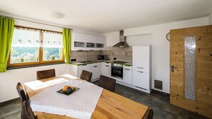 A compact kitchen with white cabinets, a stove, and a refrigerator. There's a dining table with chairs and a view of a mountainous landscape through a window. - Apartment Weisse Zinne (St.Johann)