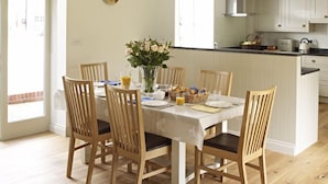 A dining area with a wooden table and chairs, a vase of flowers, and a kitchen in the background. - Manor Farm Courtyard Cottages (Thatcham)