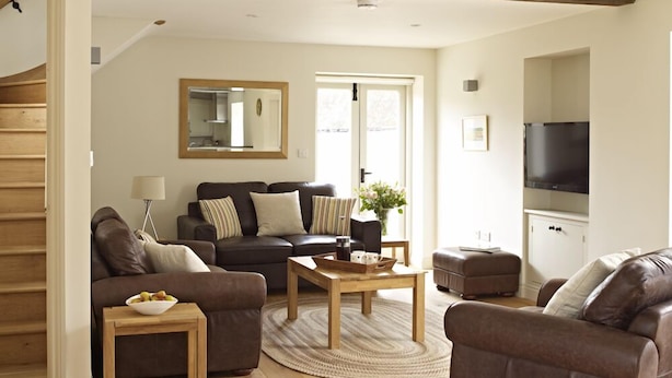 A modern living room with a sofa, coffee table, and ottoman, wooden flooring, and a television. - Manor Farm Courtyard Cottages (Thatcham)