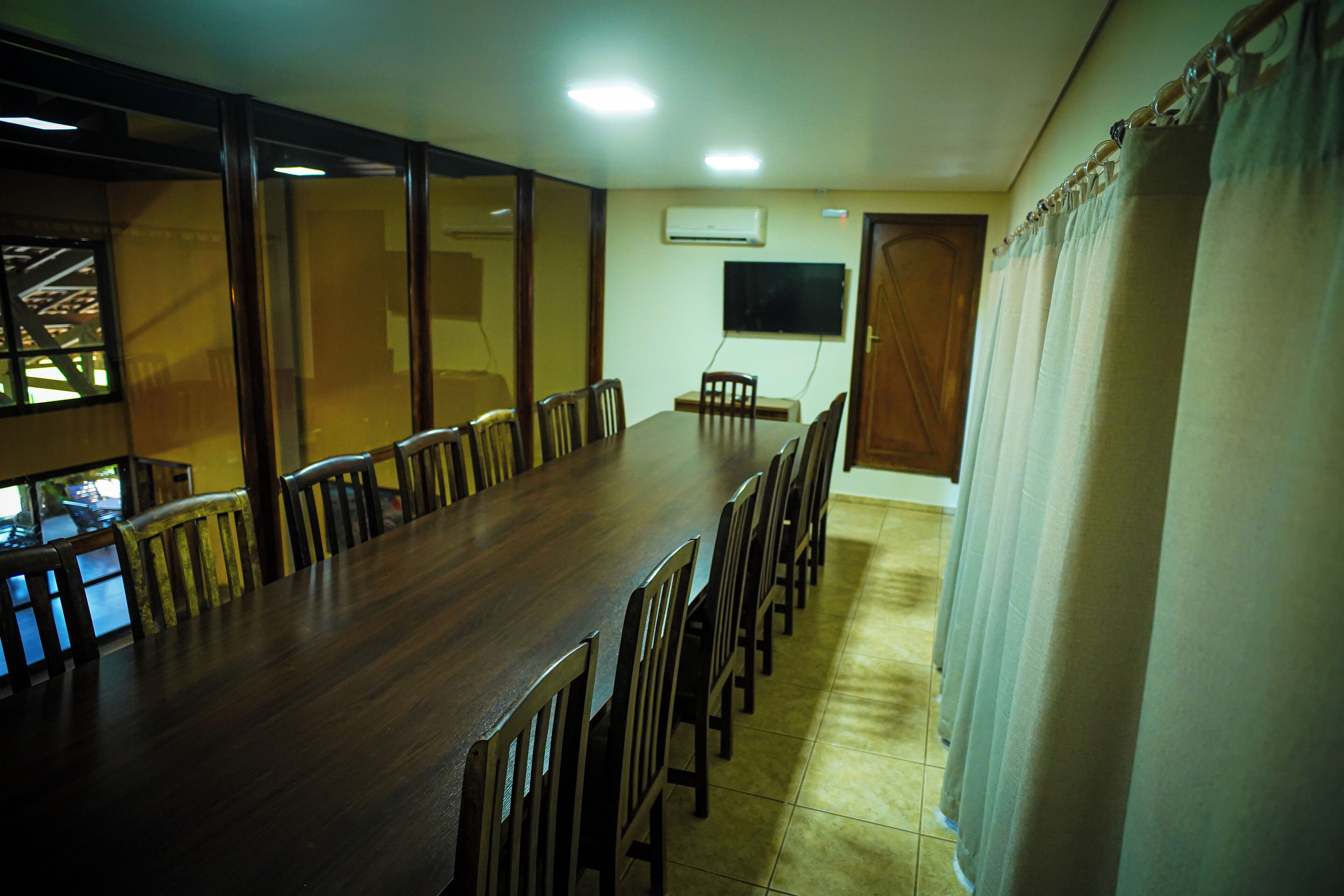 A long dining table with chairs, a television on the wall, and a window with a view of a building.