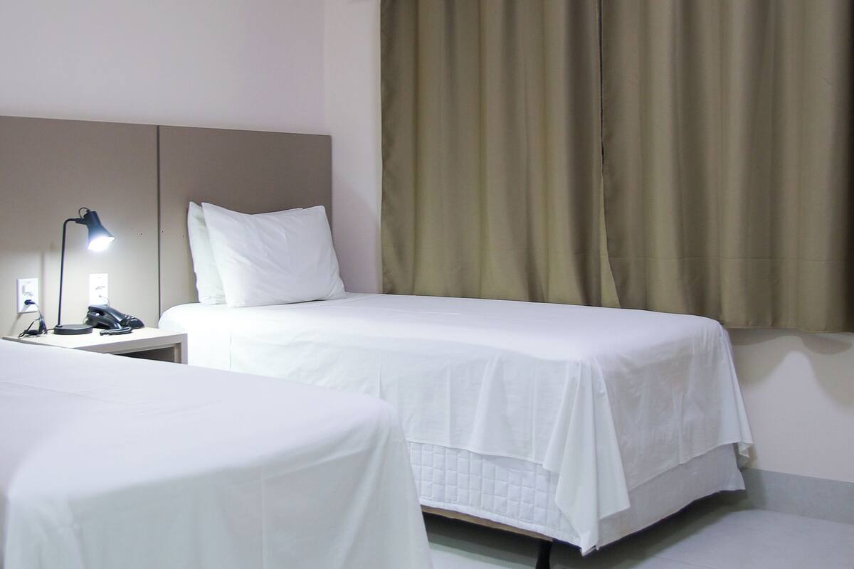 A hotel room with two beds, a nightstand, a lamp, and a window with curtains.